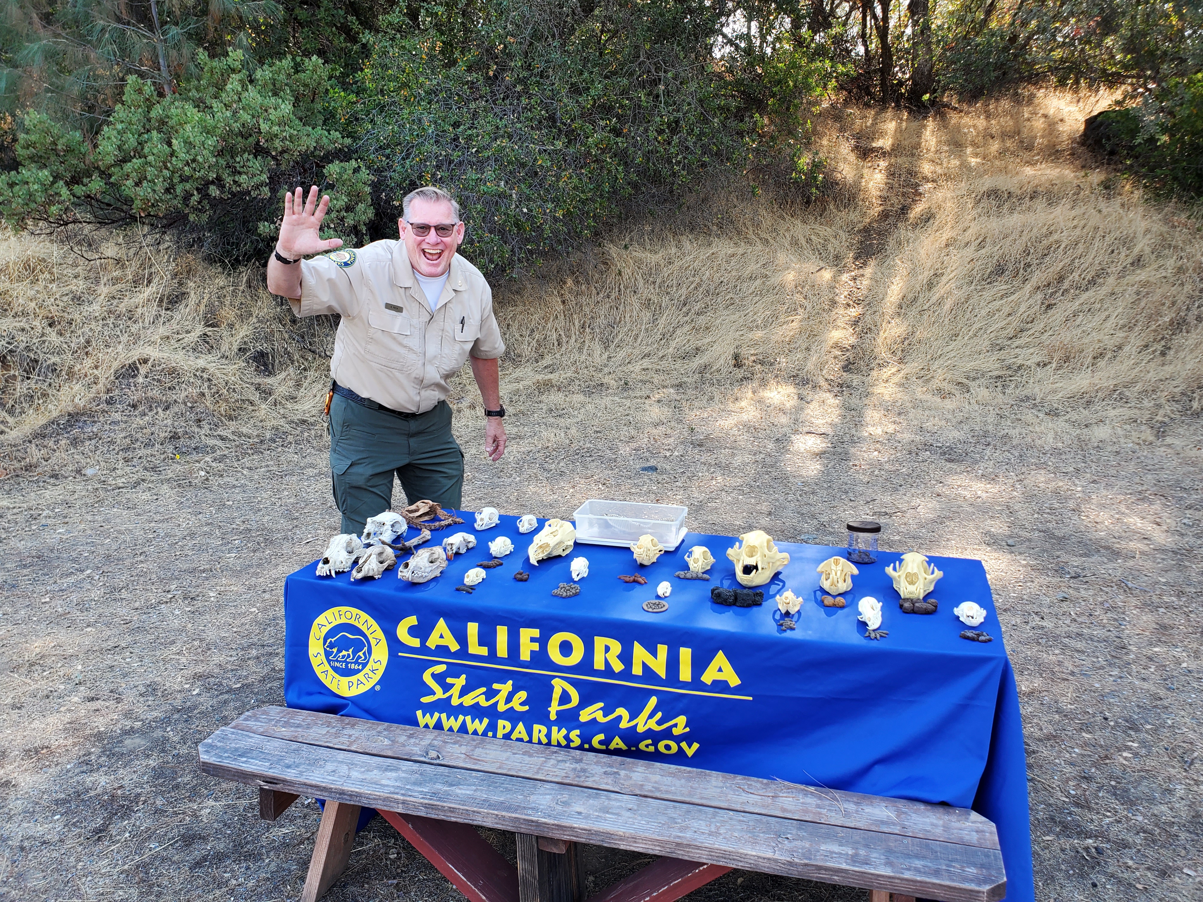 Tall man in park uniform smiling and waving next to a table with scat and skulls